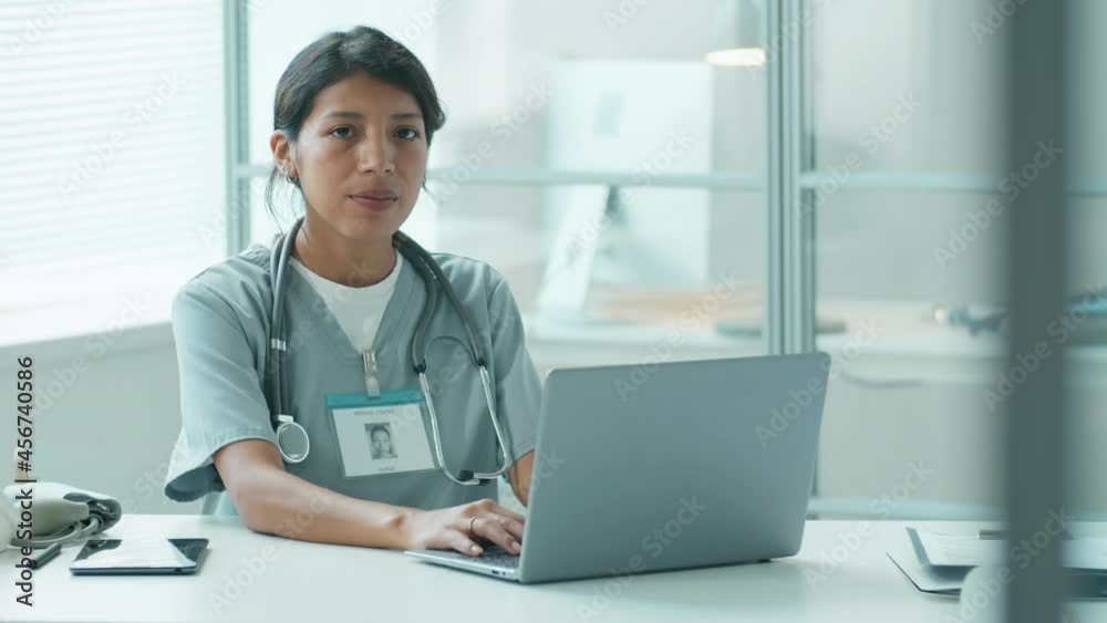 Beautiful Hispanic female doctor typing on laptop and then smiling at camera while working in medical office Beautiful Hispanic female doctor typing on laptop and then smiling at camera while working in medical office