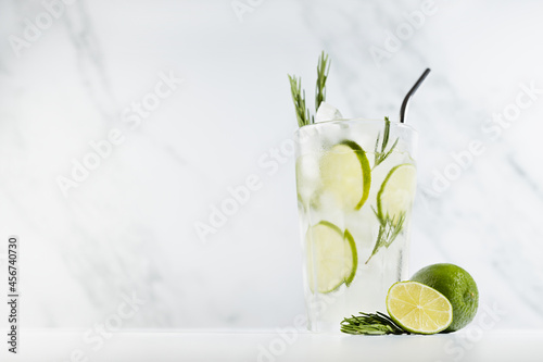 Healthy summer vitamin cold tonic drink with lime juice, ice, green rosemary twig, silver straw, ingredients on white wood table and soft light marble wall.