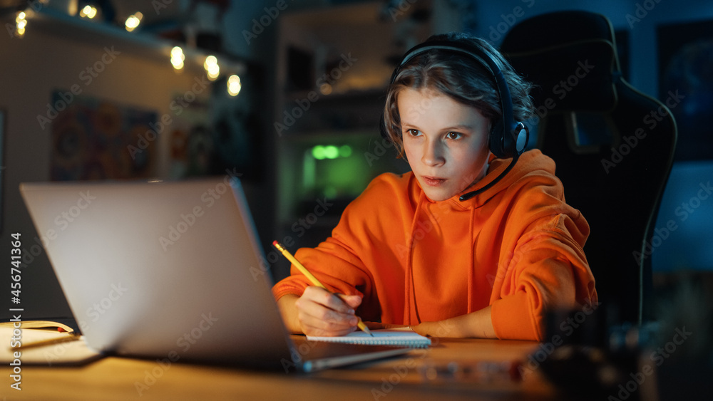 Smart Young Boy in Headphones Attending Online Class on Laptop Computer in Cozy Dark Room at Home. Happy Teenager Browsing Educational Research Online, Writing in Notebook, Studying School Homework. Smart Young Boy in Headphones Attending Online Class on Laptop Computer in Cozy Dark Room at Home. Happy Teenager Browsing Educational Research Online, Writing in Notebook, Studying School Homework.