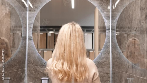 A blonde woman stands with her back to the camera and turns on the illumination of the mirror that hangs in the bathroom