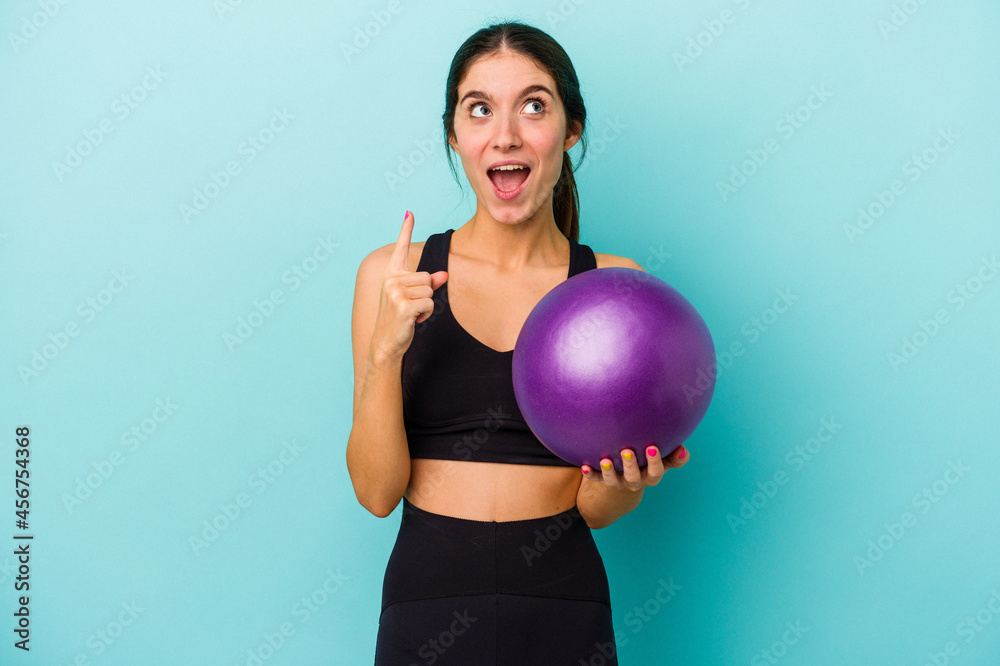 Young caucasian fitness woman holding a ball isolated on blue background pointing upside with opened mouth. Young caucasian fitness woman holding a ball isolated on blue background pointing upside with opened mouth.