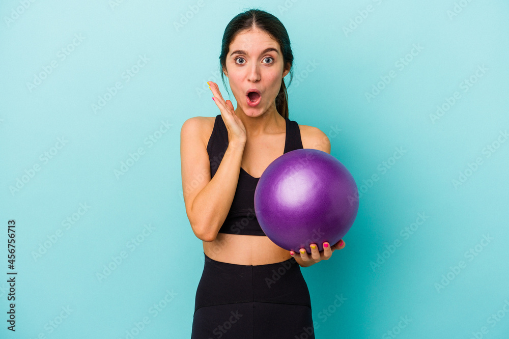 Young caucasian fitness woman holding a ball isolated on blue background surprised and shocked. Young caucasian fitness woman holding a ball isolated on blue background surprised and shocked.