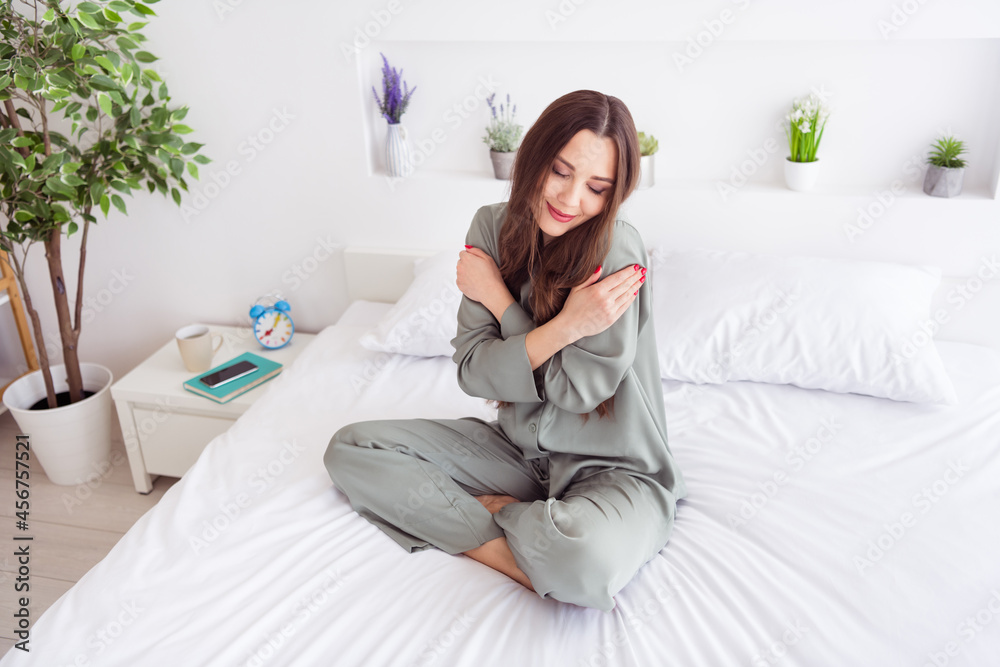 Photo of shiny charming young woman grey nightwear sitting bed hugging herself smiling inside indoors home room