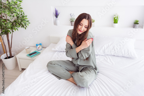 Photo of shiny charming young woman grey nightwear sitting bed hugging herself smiling inside indoors home room
