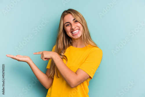 Young caucasian blonde woman isolated on blue background  excited holding a copy space on palm.