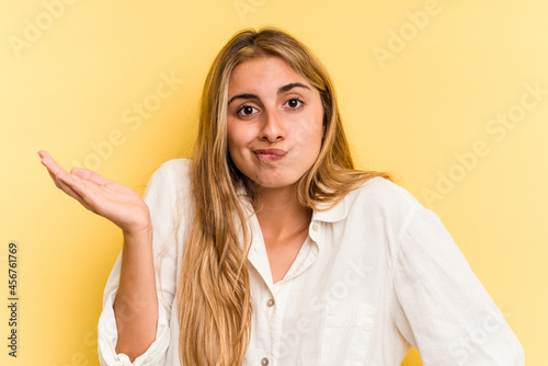 Young caucasian blonde woman isolated on yellow background showing a copy space on a palm and holding another hand on waist.