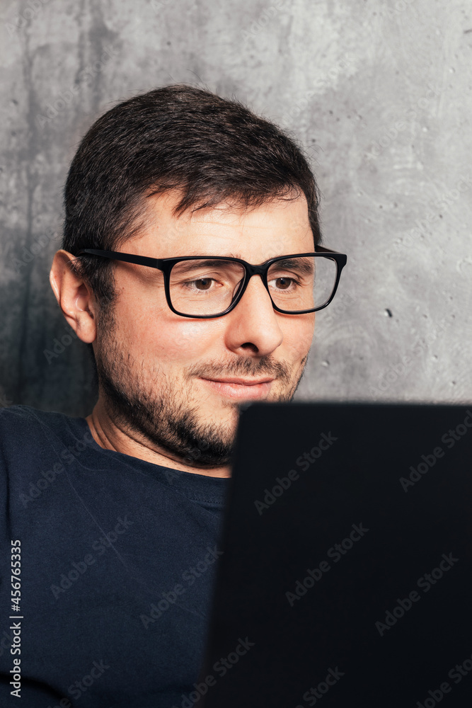 Handsome bearded man in glasses looking at his laptop screen against background of concrete wall in modern office. Closeup view Handsome bearded man in glasses looking at his laptop screen against background of concrete wall in modern office. Closeup view
