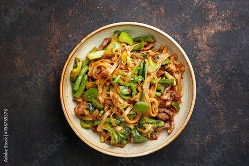 Oriental noodles, top view. Beef fried on a wok with rice noodles, green beans, zucchini, pak choi and green peppers in spicy soy-oyster sauce, in a bowl, on black stony background. Thai cousine dish.