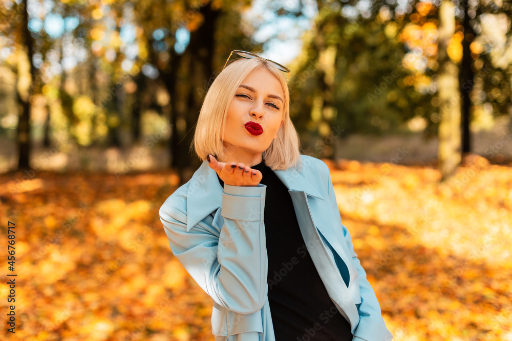 Pretty happy woman with red lips in fashionable clothes sends an air kiss on the background of colorful golden autumn foliage on a sunny holidays. Pretty happy woman with red lips in fashionable clothes sends an air kiss on the background of colorful golden autumn foliage on a sunny holidays.