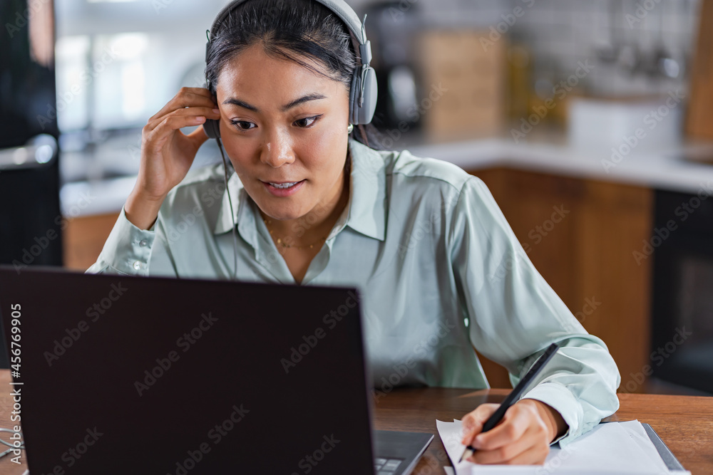 Fototapeta premium Young Asian woman attending online foreign language classes. Sitting in front of laptop computer with headphones listening course and taking notes.