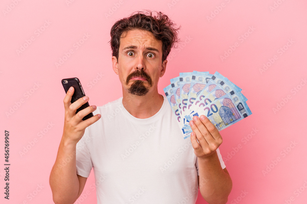 Young caucasian man holding bills and piggy bank isolated on pink background Young caucasian man holding bills and piggy bank isolated on pink background