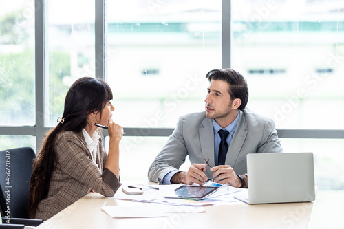 Businessman and businesswoman sharing laptop while sitting at a desk in office building room.