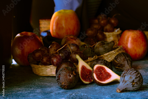 Still life of figs, grapes and apples on wicker baskets, a blue cloth and a dark background.