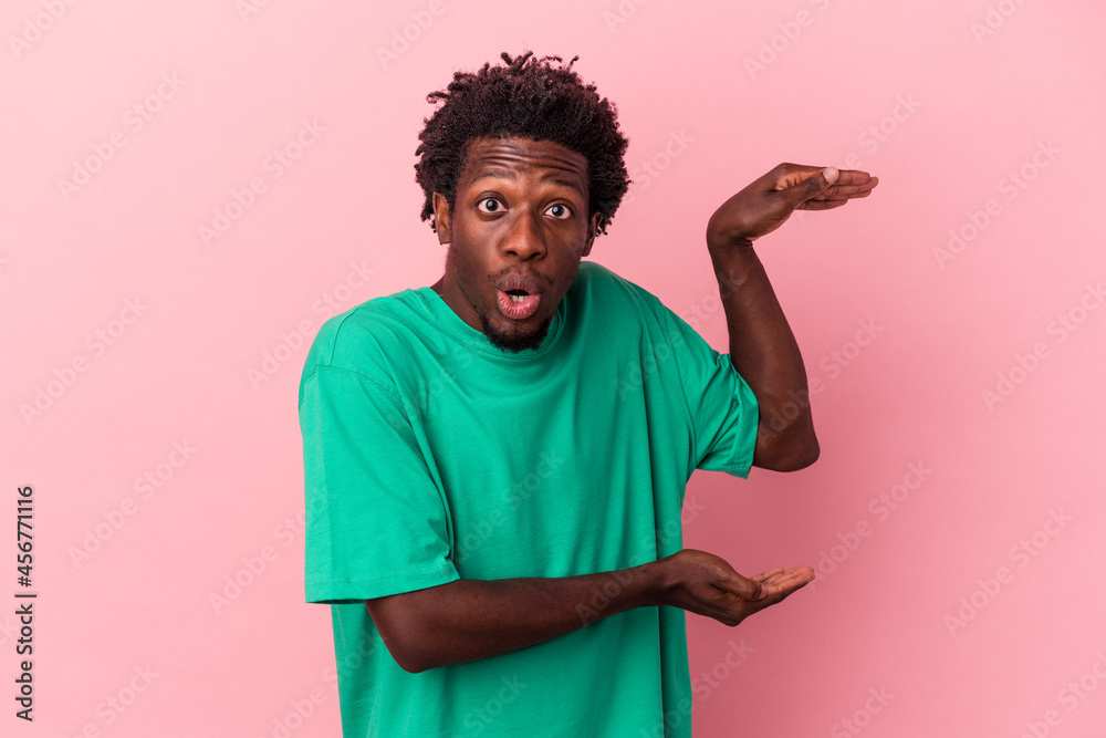 Young african american man isolated on pink background shocked and amazed holding a copy space between hands.