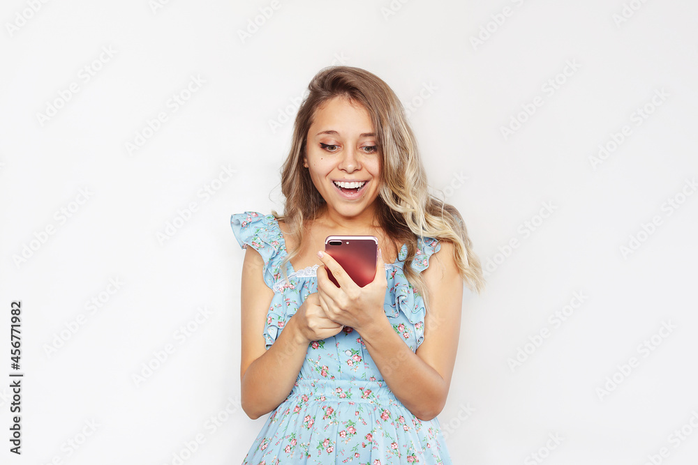 A young caucasian amazed smiling blonde woman with wavy hair in a blue dress smiles holding a mobile phone looking at the screen isolated on a white background. The amazed girl uses her smartphone A young caucasian amazed smiling blonde woman with wavy hair in a blue dress smiles holding a mobile phone looking at the screen isolated on a white background. The amazed girl uses her smartphone