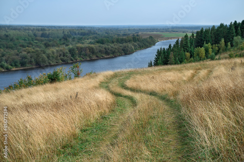 A winding road in the middle of the field, leading down to the river. The nature of the Northern Urals (Russia) with mountains, the Kolva River, tall yellow grass and a forest in the distance. Peace 