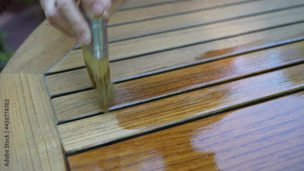 Closeup of paintbrush applying varnish to teak table furniture outside.