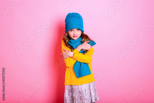Cold. Virus. Blonde kid child girl is dressed in a warm sweater, scarf, and hat on a pink background. Winter and autumn seasons.