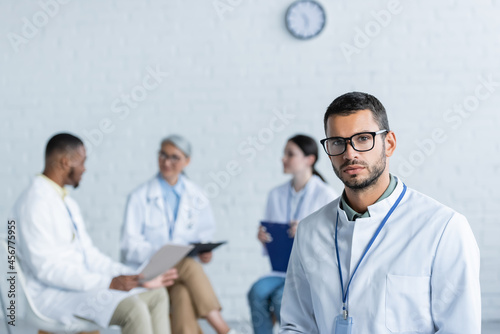 young physician in eyeglasses looking at camera while multiethnic doctors talking on blurred background