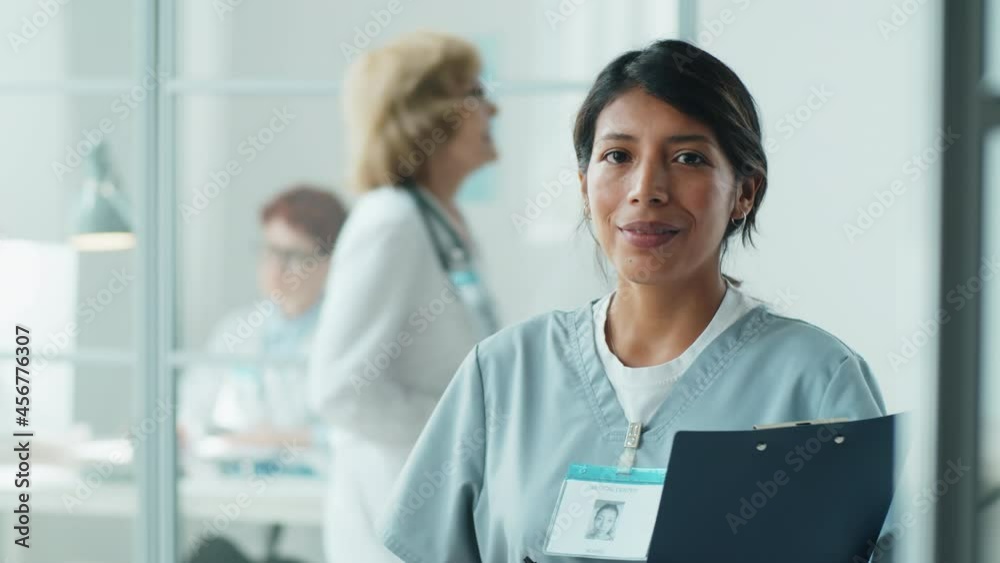 Portrait of cheerful Hispanic nurse in medical uniform holding clipboard, looking at camera and smiling while standing in clinic Portrait of cheerful Hispanic nurse in medical uniform holding clipboard, looking at camera and smiling while standing in clinic
