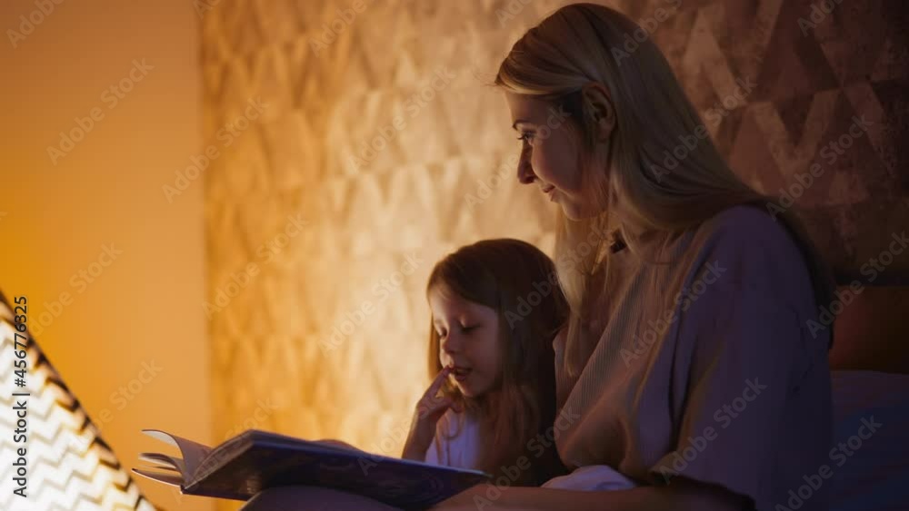 Little child girl touches lip with finger and reads book sitting near positive dimly lit mom on bed against play teepee in shaded bedroom closeup slow motion