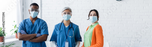 multiethnic doctors and young woman in medical masks looking at camera in vaccination center, banner