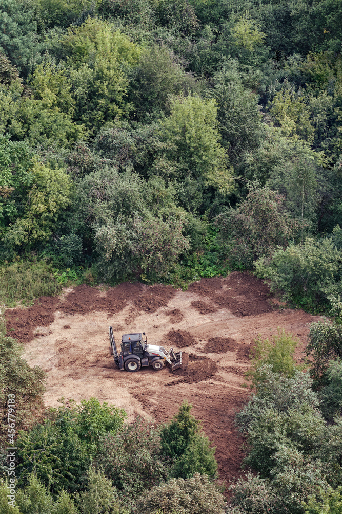 The excavator prepares the soil on the field in the forest among the trees for landscaping and agriculture, top view