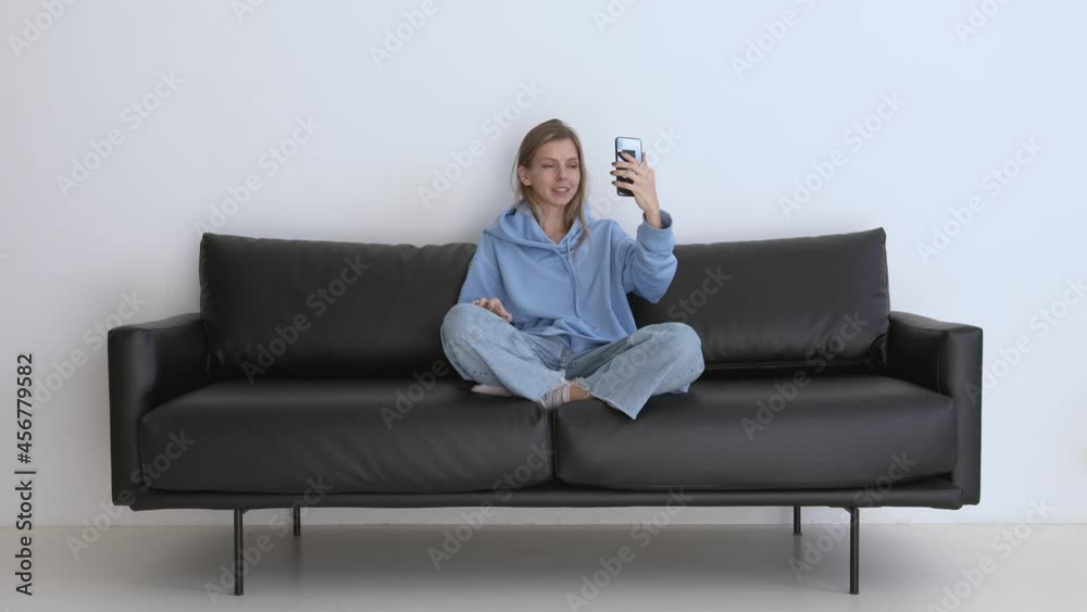 Smiling and cheerful woman having a video call with mobile phone in hand, in casual clothes with legs on a couch. Caucasian woman giving a talk on a video camera at home interior Smiling and cheerful woman having a video call with mobile phone in hand, in casual clothes with legs on a couch. Caucasian woman giving a talk on a video camera at home interior