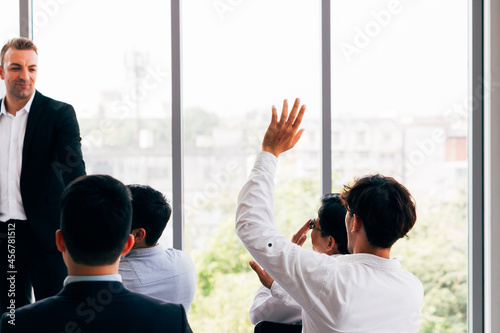 Back view of guy in shirt lifting hand and asking question while sitting amidst colleagues during business conference in office