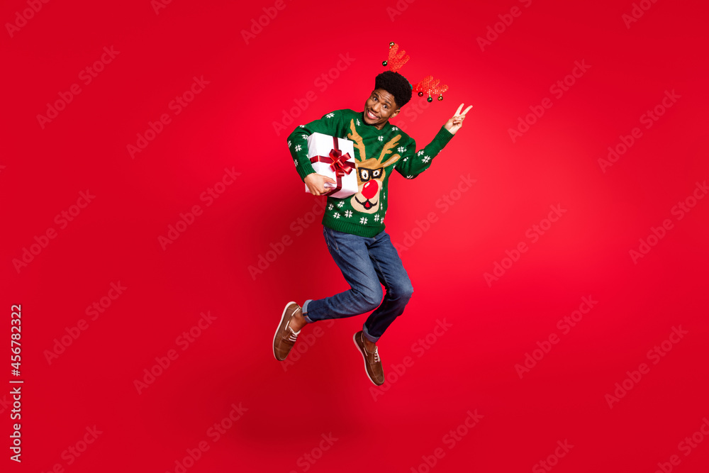 Full length body size view of trendy handsome funky cheery guy jumping holding gift showing v-sign isolated over vivid red color background