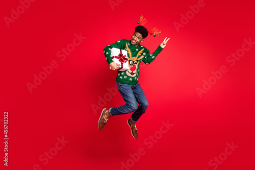 Full length body size view of trendy handsome funky cheery guy jumping holding gift showing v-sign isolated over vivid red color background
