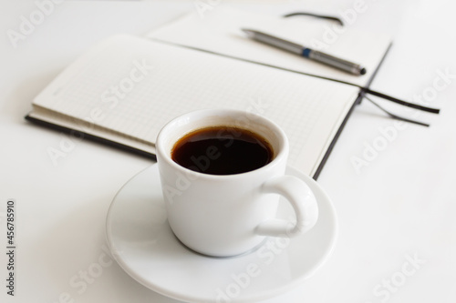 The concept of a workplace with coffee in a neat white mug on a saucer and an open notebook with a pen on a modern white office desk. Break for a delicious and hot coffee. Minimalism. Side view