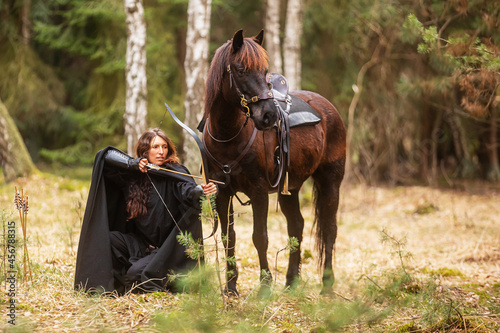 Attractive woman in fantasy costume draw bow and arrow next to horse