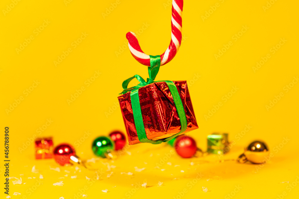 Candy cane lollipop holding a gift box with a Christmas present on a yellow background, Christmas sweets with new year decorations