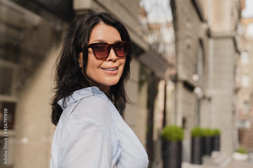 Confident middle-aged woman in sunglasses smiling while looking at the camera during walk in city. Mature businesswoman mother wife in sun eyewear outdoors.