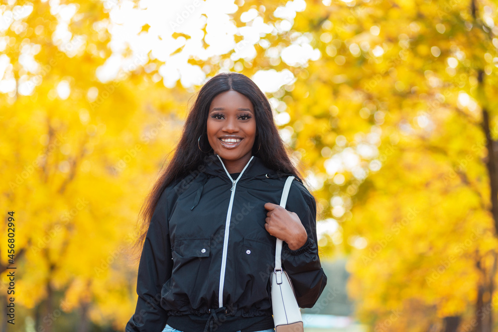 Beautiful happy black smiling young woman in fashionable casual clothes with a jacket, jeans and a handbag walks in an autumn park outdoors. Girl on a background of bright yellow foliage Beautiful happy black smiling young woman in fashionable casual clothes with a jacket, jeans and a handbag walks in an autumn park outdoors. Girl on a background of bright yellow foliage