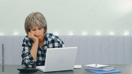 An elderly tired woman with glasses sits at her laptop while working online at home, then almost falls asleep and wakes up. An elderly woman can hardly overcome fatigue.
