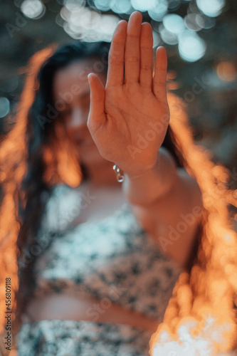 Muchacha joven sonriente y feliz de pelo rizado evitando una fotografia haciendo la señal de parar o stop junto a la naturaleza de los arboles y la puesta de sol por la mañana