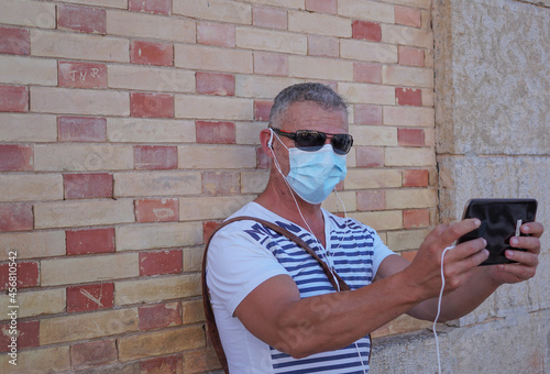 Hombre de mediana edad con mascarilla sanitaria de vacaciones en Altea con Tablet en video conferencia y gafas de sol photo