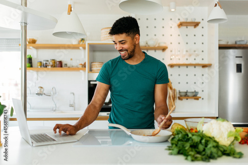 A young, happy African man is standing in the kitchen, looking at a recipe on a laptop and preparing a healthy dinner.