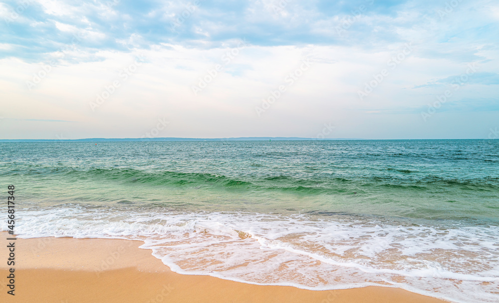 Tropical turquoise-colored seawater and white waves on the beach. White clouds on the soft blue sky in the background.