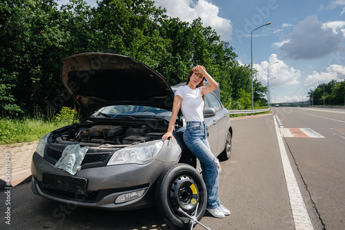 A young girl stands near a broken car with a broken wheel in the middle of the highway and is frustrated waiting for help on a hot day. Breakdown and breakdown of the car. Waiting for help. © Andrii