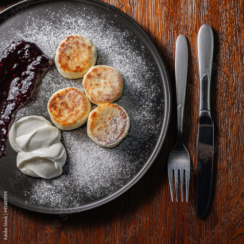 Overhead view of plate with appetizing traditional Russian cheese pancakes with sour cream and berry jam sprinkled with sugar powder on wooden table in cafe 