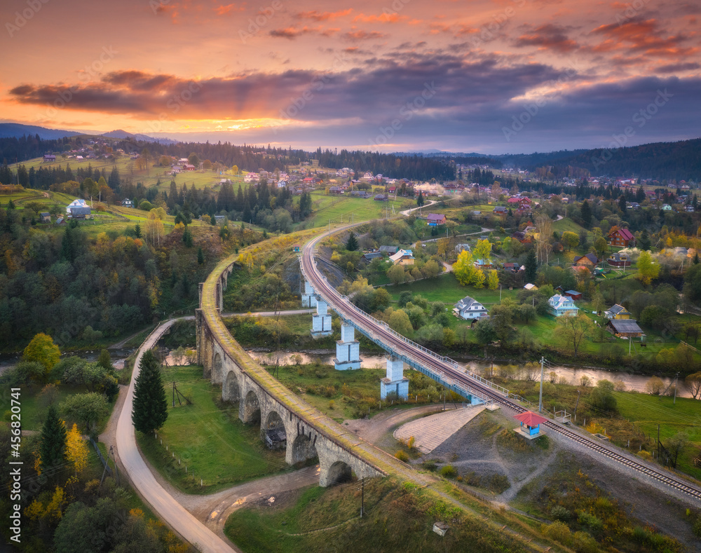 Beautiful old viaduct at sunset in carpathian mountains in autumn in Ukraine. Aerial view of railway bridges, railroad, river, green meadows, trees, houses, hills and colorful sky in fall. Top view