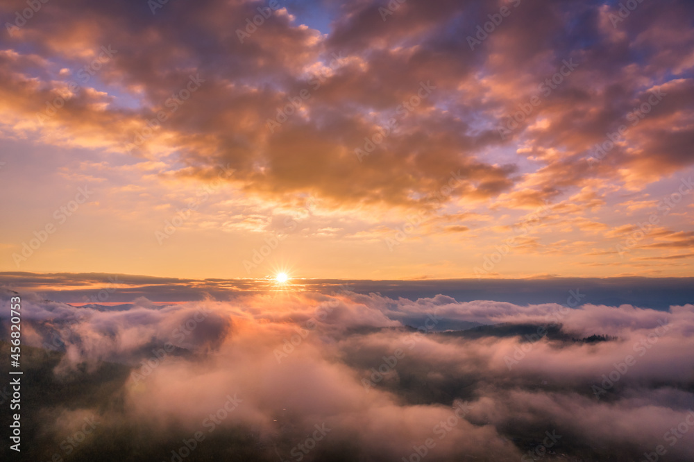 Mountains in low clouds at sunrise in summer. Aerial view of mountain peak in fog. Beautiful landscape with high rocks, forest, colorful sky. View from above of mountain valley in clouds. Foggy hills	