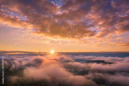 Mountains in low clouds at sunrise in summer. Aerial view of mountain peak in fog. Beautiful landscape with high rocks, forest, colorful sky. View from above of mountain valley in clouds. Foggy hills 