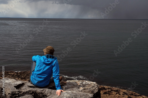 Man in blue jacket and a hat sitting on a rock by the ocean. Back to camera. Storm clouds over water surface. Dark depressive colors. Loneliness concept. West coast of Ireland.