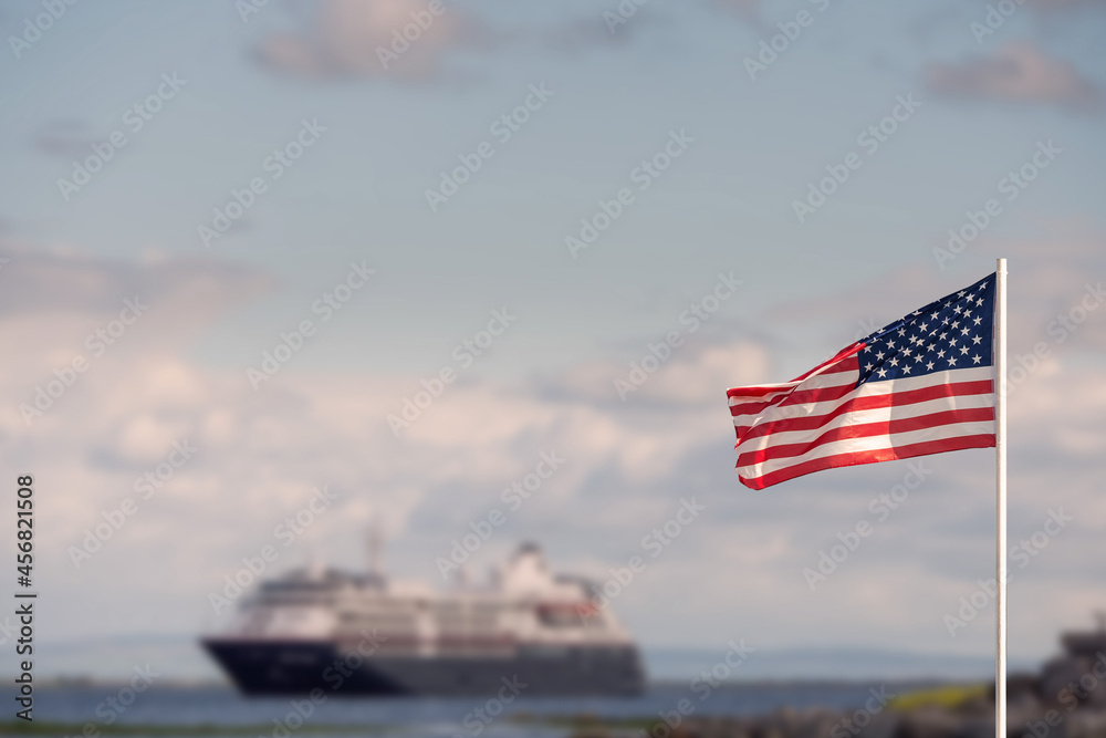 Waving flag of United States of America against pastel light blue color sky in focus. Cruise ship out of focus in the background. Abstract holiday and travel concept