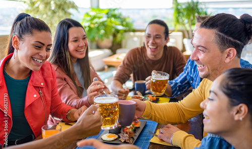 multiethnic group of friends sitting on a table in a bar restaurant making a toast with beers and drinks. diverse people celerating brunch together enjoying happy holidays. lifestyle and joy concept