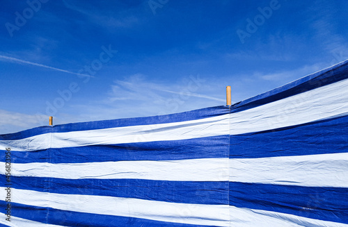 Windbreak beach windscreen in blue and white stripes against blue sky with mild clouds in the background.